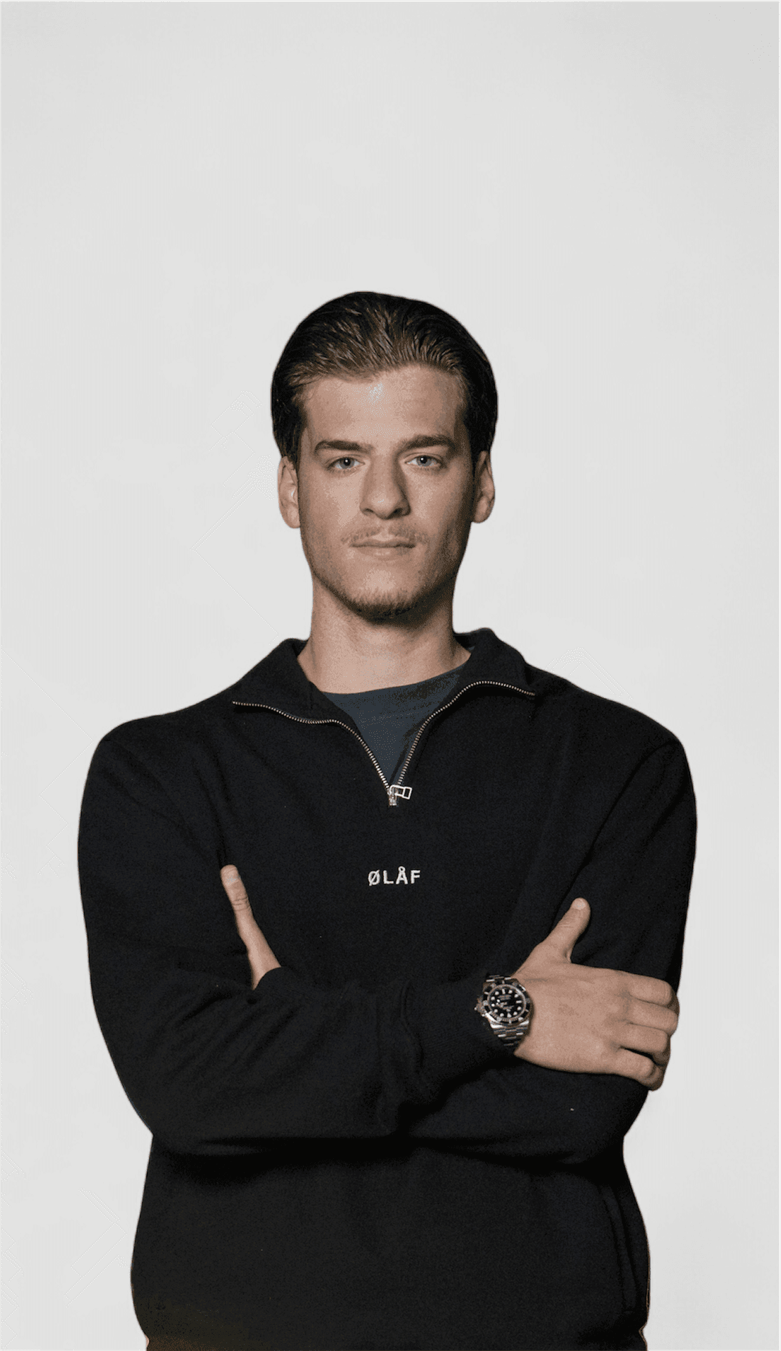Man with crossed arms wearing a black sweatshirt and watch, standing against a plain background.
