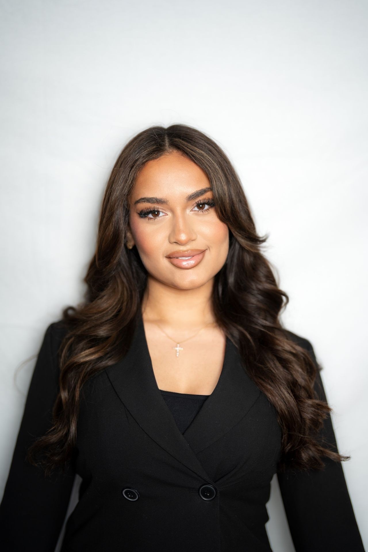 Woman with long brown hair wearing a black blazer and necklace, posing against a plain background.